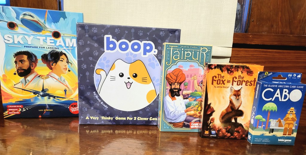 Five game boxes sitting on a table, in descending hight order from left to right. They are: Sky Team, boop, Jaipur, The Fox In The Forest, and Cabo