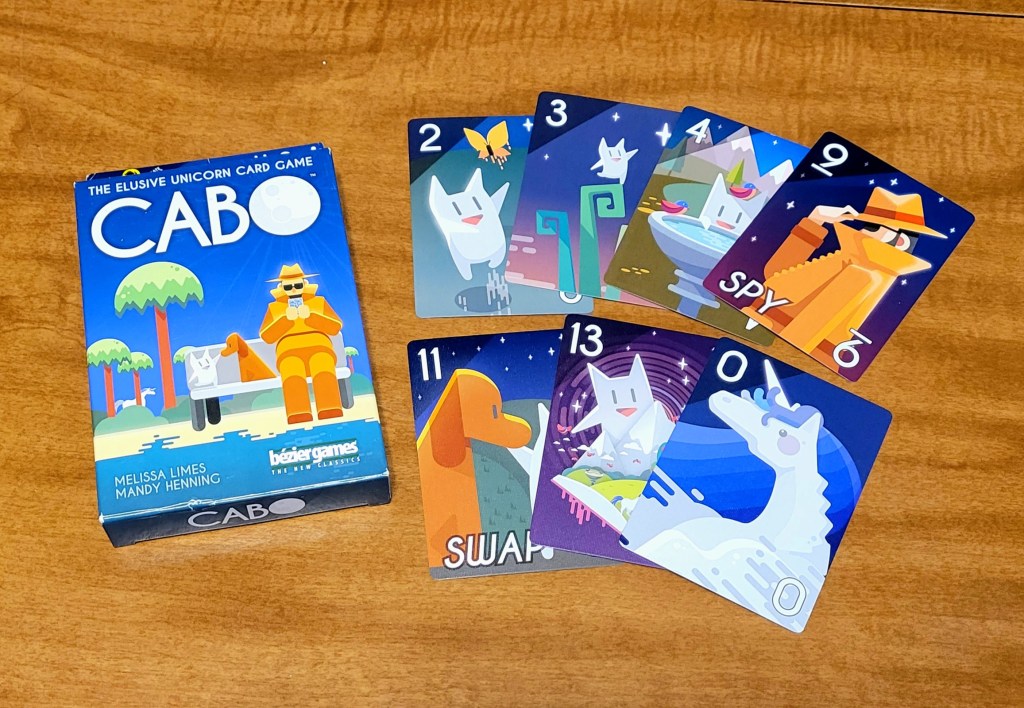 The game Cabo laid out on a table. The box is a deep blue and reads, 'The Elusive Unicorn Card Game CABO' with an illustration of a person, dog and cat on a bence, with trees, bushes, and a unicorn in the background. To the right of the box are seven cards, each with a number in the top left corner. The cards also contain illustrations of the cat, dog, person, and unicorn from the box engaged in various activities.