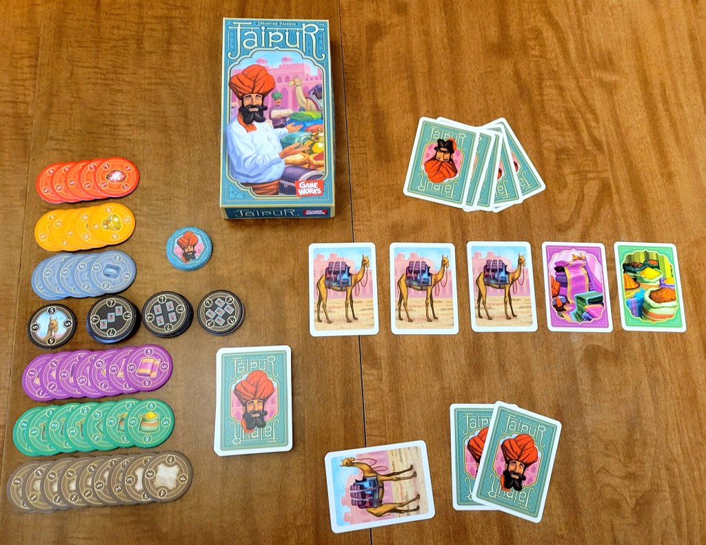 The elements of the game Jaipur spread out on a table. The box has the word Jaipur in an ornate font, a digital painting of a bearded man with a white shirt and tunic, a number of brightly colored, valuable looking objects. The game consists of stacks of tokens in orange, yellow, blue, black, purple, green and brown, and cards. some of the cards show the bearded man's face and the word "Jaipur." Others show camels, cloth, or sacks of foodstuffs.