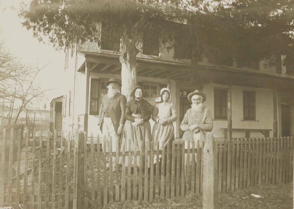 ID: Aged, sepia-toned photograph of four people in 19th century clothing standing behind a picket fence but in front of a white house, under a tree.  All have light skin.  The two on the ends are men with shite beards and broad-brimmed hats.  The two in the middle wear dresses and bonnets.  Their expressions are difficult to make out,  