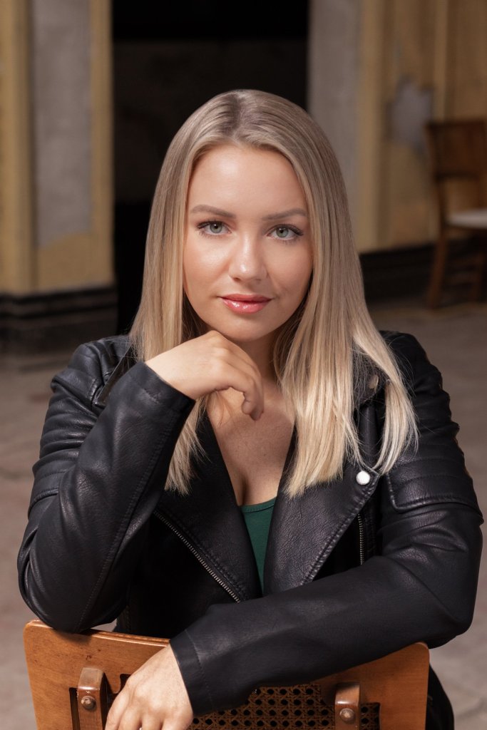 A white person with straight blonde hair and elegant makeup sits backward on a wooden chair. They are wearing a black leather jacket. On hand is resting on the back of the chair and the other is raised to their chin.