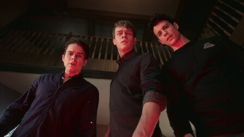 Three young white men in a darkened room stare down at the camera with concerned expressions.  