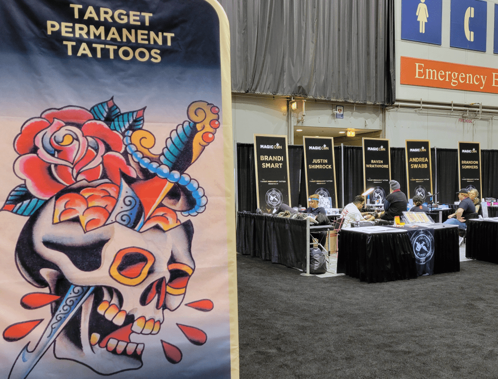 Photo showing the "Target Permanent Tattoos" sign next to a section of tables, chairs and signs for various tattoo artists giving tattoos to con attendees.