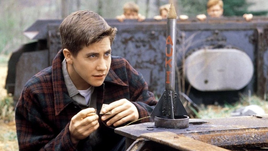 Disappointingly unflattering photo of a young Jake Gyllenhaal sitting at a work bench in front of a small metal rocket painted with the letters "AUK I."  He has light skin, short dark brown hair that flips up in the front.  He is wearing a plaid flannel shirt open at the collar, with two more shirts visible underneath.  He is hunched over the work bench but looking up with a determined expression.  