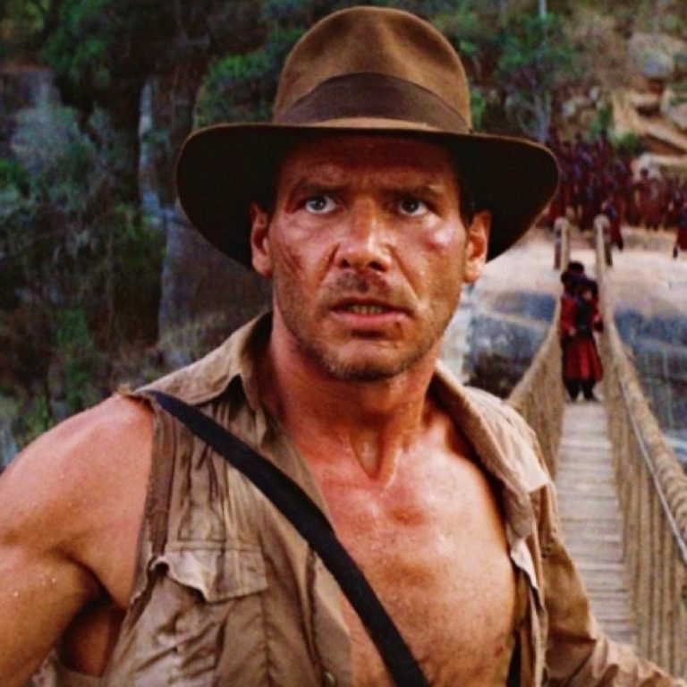 Older photo of a light skinned man with a brown fedora hat standing on a rope bridge.  He wears a brown fedora hat and a beige vest open in front, exposing defined muscles on his arms and chest.  A black sash crosses his chest diagonally.  His face is stubbly and has an intense expression.  
