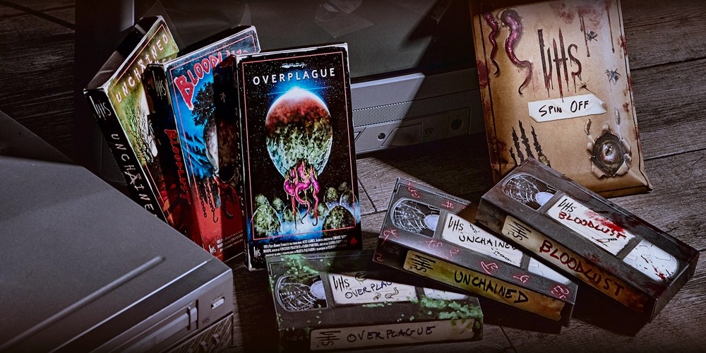 Photo of several boxes designed to look like weathered VHS tape cartridges and their boxes, staged with VCR machines.  The "boxes" shown upright on the left, read "Uncharted," "Bloodlust," and "Overplague" respectively and include spooky art in a vintage pulp horror style.  The three "tapes" are arranged horizontally in a toppled stack and are labeled with the same titles.  In the upper right there is a light brown package decorated in horror themed art and labeled "VHS Spin Off."